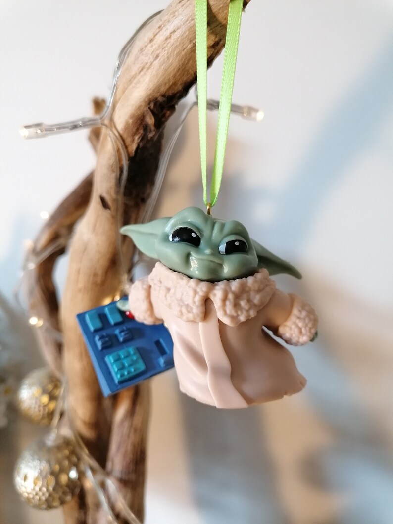 May include: A green and brown character from the Star Wars franchise, known as "The Child" or "Baby Yoda", is wearing a tan coat with a fur collar and holding a blue and silver device. The character is hanging from a branch with a green ribbon.