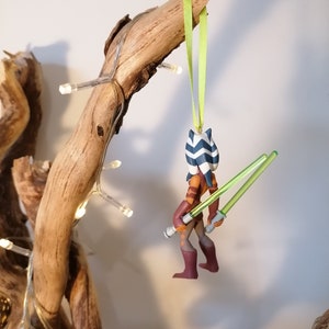 May include: A Star Wars themed Christmas ornament featuring Ahsoka Tano, a Togruta Jedi Padawan, holding two green lightsabers. The ornament is made of plastic and is painted in shades of brown, blue, and white.