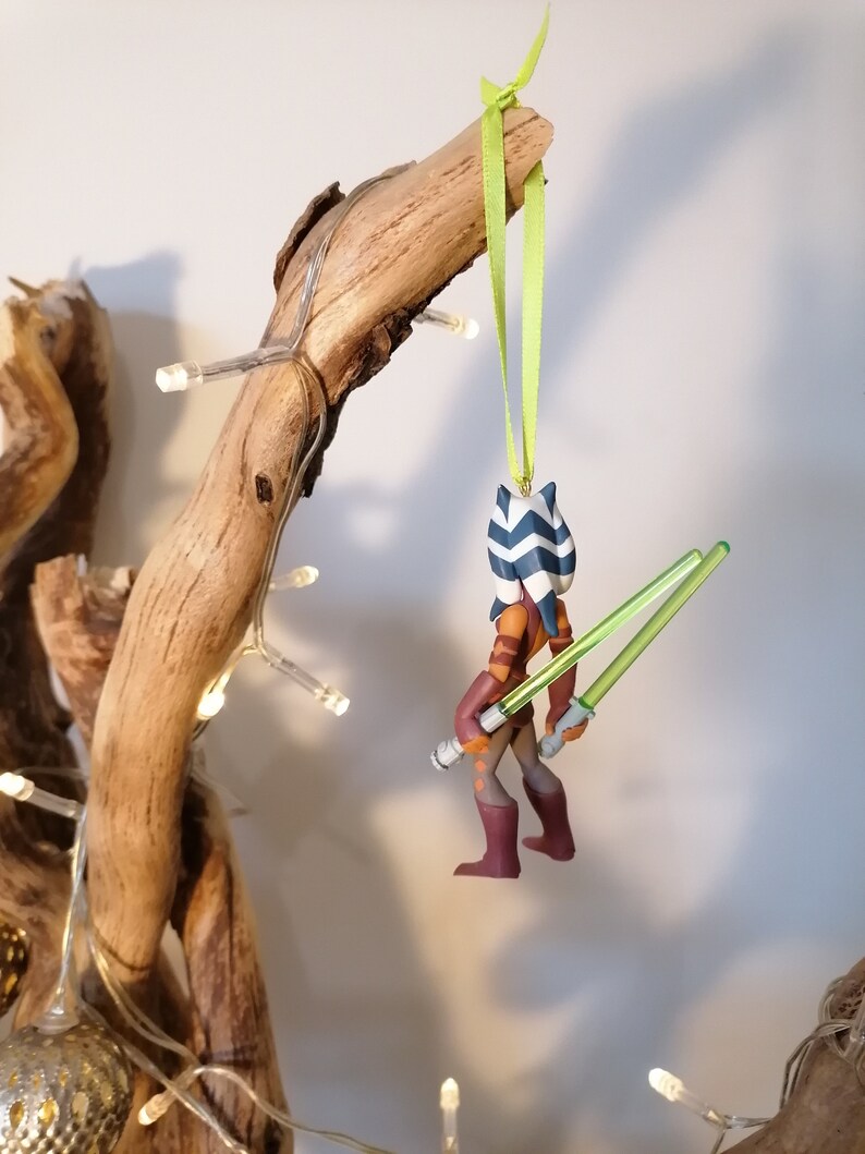 May include: A Star Wars themed Christmas ornament featuring Ahsoka Tano, a Togruta Jedi Padawan, holding two green lightsabers. The ornament is made of plastic and is painted in shades of brown, blue, and white.