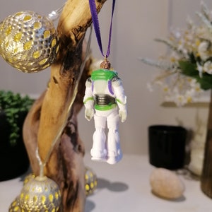 May include: A white and green plastic toy astronaut ornament with a purple ribbon hanging from a wooden branch. The astronaut is wearing a space suit with a black belt and a clear visor.