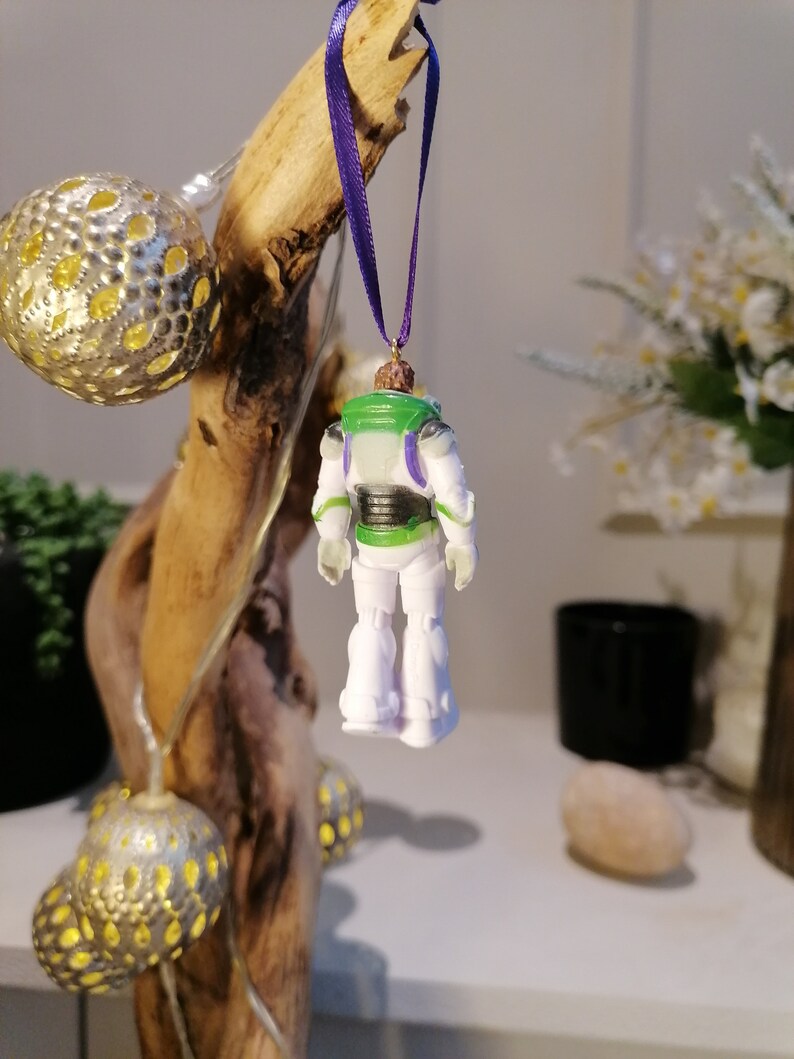 May include: A white and green plastic toy astronaut ornament with a purple ribbon hanging from a wooden branch. The astronaut is wearing a space suit with a black belt and a clear visor.