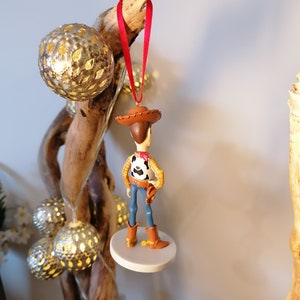 Op de afbeelding: Een rood en wit cowboy-figuurtje ornament van Woody uit Toy Story. De ornament hangt aan een rood lint en staat op een witte basis.