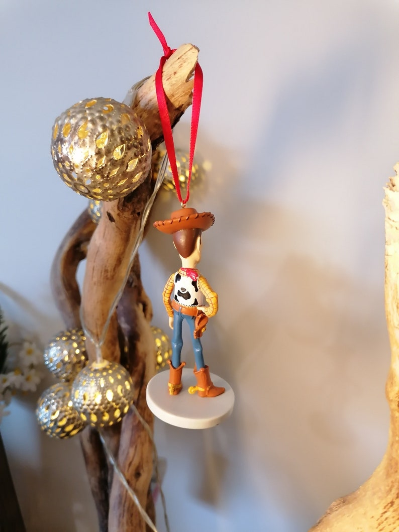 Op de afbeelding: Een rood en wit cowboy-figuurtje ornament van Woody uit Toy Story. De ornament hangt aan een rood lint en staat op een witte basis.
