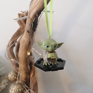 Puede incluir: Un adorno de Yoda verde colgando de una rama con una cinta verde. El adorno est&aacute; sobre una base hexagonal negra.