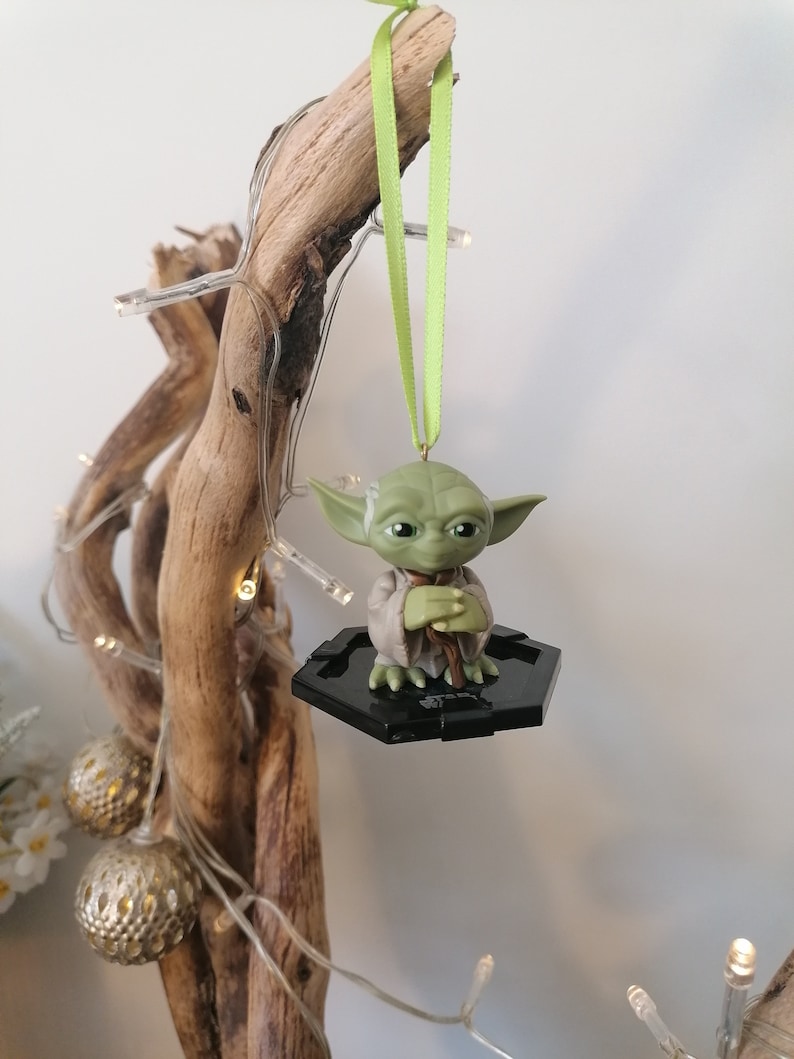 Puede incluir: Un adorno de Yoda verde colgando de una rama con una cinta verde. El adorno est&aacute; sobre una base hexagonal negra.