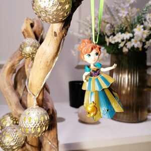 May include: A plastic ornament of a red-haired princess wearing a gold and blue dress with a bird design. The ornament is hanging from a green ribbon on a branch with gold and white decorative lights.