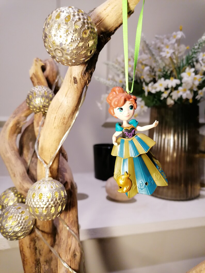 May include: A plastic ornament of a red-haired princess wearing a gold and blue dress with a bird design. The ornament is hanging from a green ribbon on a branch with gold and white decorative lights.