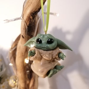 May include: A green and brown Baby Yoda Christmas ornament hanging from a green ribbon. The ornament is made of plastic and has a detailed face with large eyes.