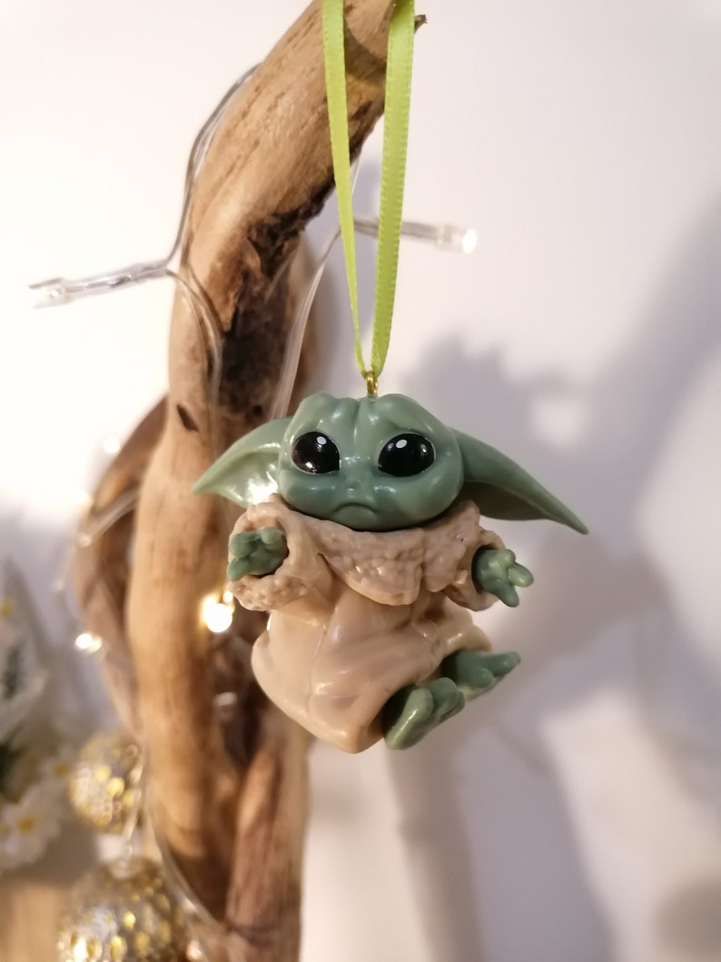 May include: A green and brown Baby Yoda Christmas ornament hanging from a green ribbon. The ornament is made of plastic and has a detailed face with large eyes.