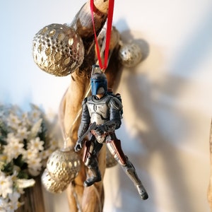 May include: A blue and gray Star Wars character ornament hanging from a branch with a red ribbon. The ornament is a depiction of Boba Fett, a bounty hunter from the Star Wars universe.