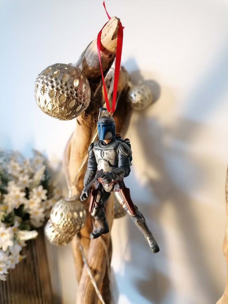 May include: A blue and gray Star Wars character ornament hanging from a branch with a red ribbon. The ornament is a depiction of Boba Fett, a bounty hunter from the Star Wars universe.
