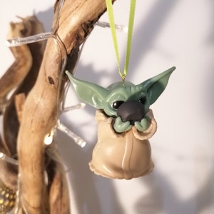 May include: A green and brown Christmas ornament shaped like a character from the Star Wars franchise. The ornament is hanging from a green ribbon.