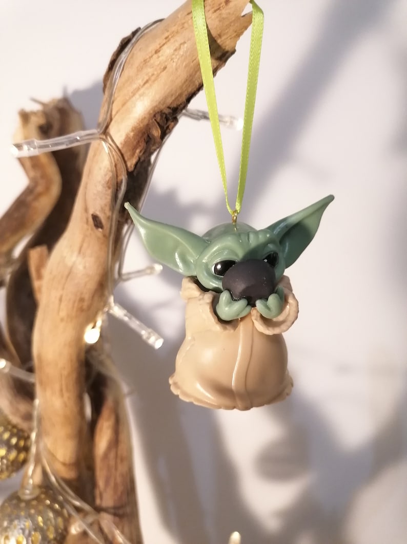 May include: A green and brown Christmas ornament shaped like a character from the Star Wars franchise. The ornament is hanging from a green ribbon.