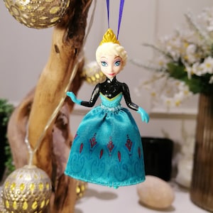 K&ouml;nnte beinhalten: Ein blauer und schwarzer Plastik-Anh&auml;nger mit der Disney-Frozen-Figur Elsa, die ein blaues und wei&szlig;es Kleid mit einer goldenen Krone tr&auml;gt. Der Anh&auml;nger h&auml;ngt an einem lila Band.