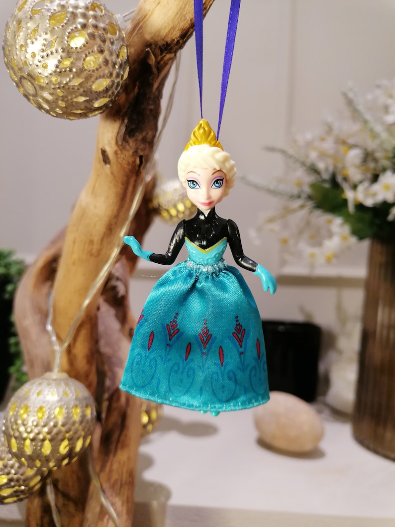 K&ouml;nnte beinhalten: Ein blauer und schwarzer Plastik-Anh&auml;nger mit der Disney-Frozen-Figur Elsa, die ein blaues und wei&szlig;es Kleid mit einer goldenen Krone tr&auml;gt. Der Anh&auml;nger h&auml;ngt an einem lila Band.