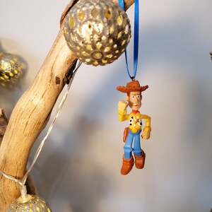 May include: A blue and white string with a plastic figurine of Woody from Toy Story hanging from it. The figurine is wearing a brown hat, a yellow shirt, blue pants, and brown boots. The string is hanging from a branch with a silver, decorative light bulb.