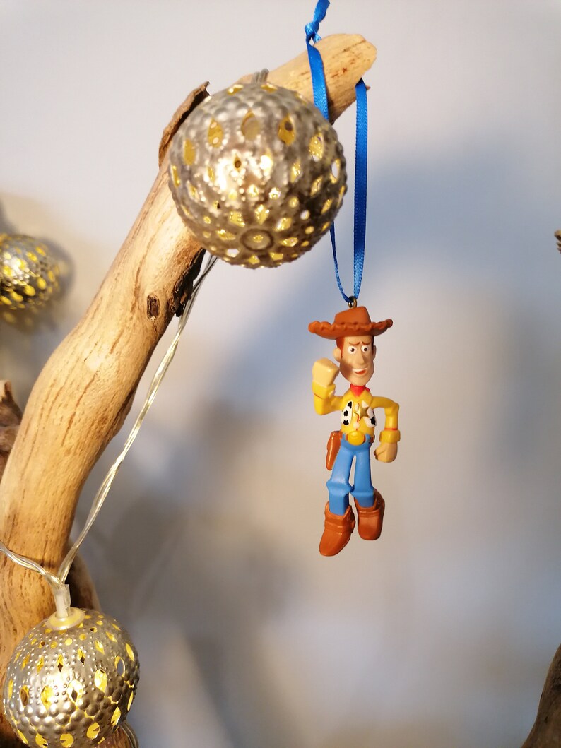 May include: A blue and white string with a plastic figurine of Woody from Toy Story hanging from it. The figurine is wearing a brown hat, a yellow shirt, blue pants, and brown boots. The string is hanging from a branch with a silver, decorative light bulb.