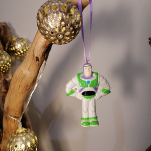 Puede incluir: Una figurita de pl&aacute;stico blanca y verde de Buzz Lightyear de la franquicia de pel&iacute;culas Toy Story colgada de una cinta morada. La figurita est&aacute; colgada de una rama con una cadena de luces.