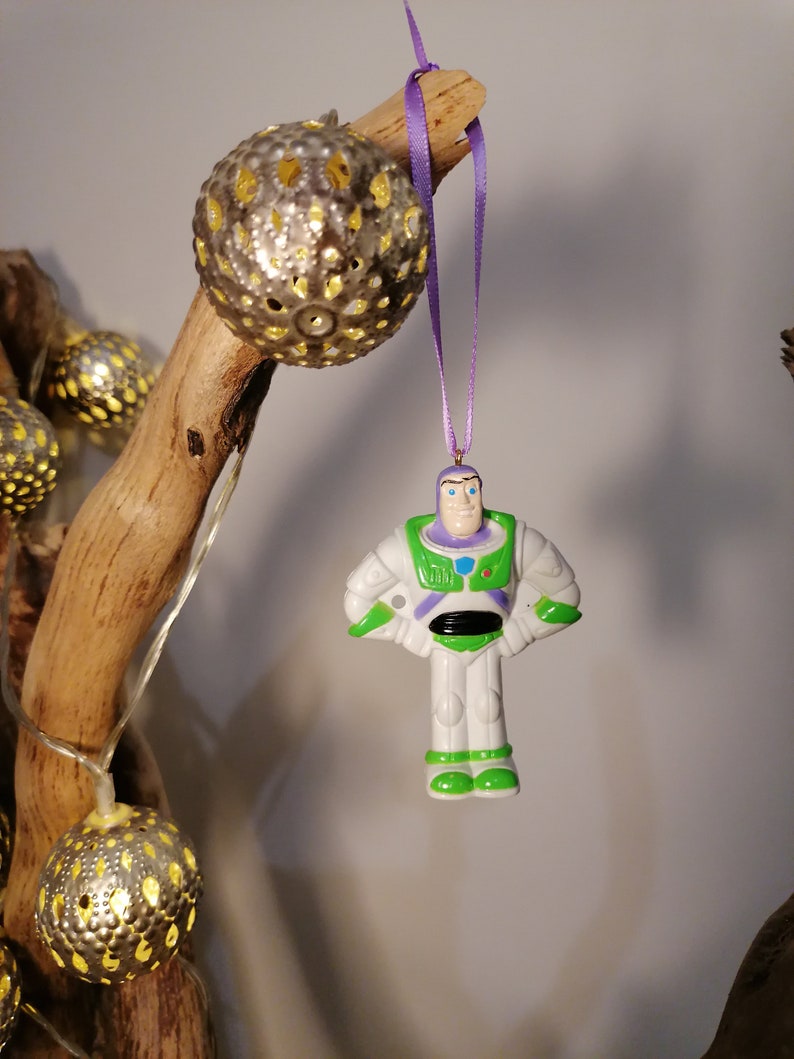 Puede incluir: Una figurita de pl&aacute;stico blanca y verde de Buzz Lightyear de la franquicia de pel&iacute;culas Toy Story colgada de una cinta morada. La figurita est&aacute; colgada de una rama con una cadena de luces.