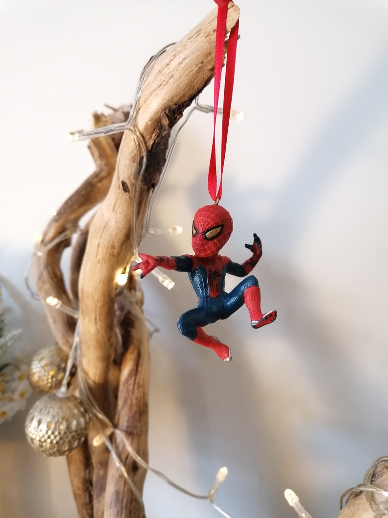 May include: A red and blue Spider-Man Christmas ornament hanging from a driftwood branch with white Christmas lights.