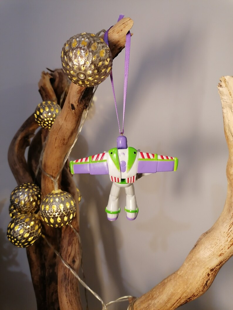 Peut inclure: Une d&eacute;coration de No&euml;l Buzz l'&Eacute;clair, suspendue par un ruban violet. L'ornement est blanc, vert et violet. Il est suspendu &agrave; un morceau de bois flott&eacute;. Des guirlandes lumineuses d&eacute;coratives avec des sph&egrave;res dor&eacute;es sont &eacute;galement dans l'image.