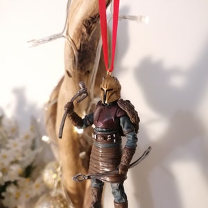 May include: A gold and brown Christmas ornament featuring a warrior figure with a red and gold helmet, holding a weapon and a bow. The ornament is hanging from a red ribbon.