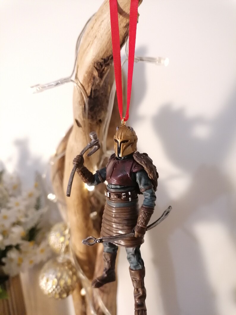 May include: A gold and brown Christmas ornament featuring a warrior figure with a red and gold helmet, holding a weapon and a bow. The ornament is hanging from a red ribbon.