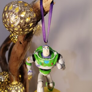 K&ouml;nnte beinhalten: Ein gr&uuml;ner und wei&szlig;er Plastik-Anh&auml;nger von Buzz Lightyear aus der Toy Story-Reihe. Der Anh&auml;nger h&auml;ngt an einem lila Band und hat eine gelbe Basis.