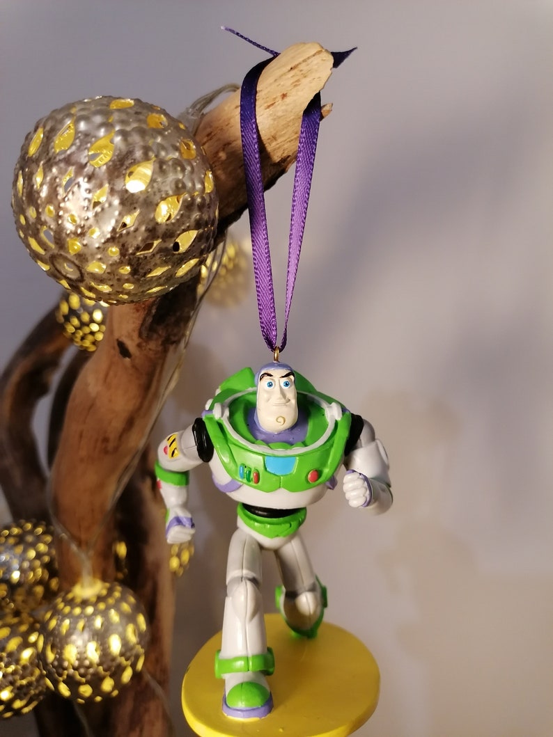 K&ouml;nnte beinhalten: Ein gr&uuml;ner und wei&szlig;er Plastik-Anh&auml;nger von Buzz Lightyear aus der Toy Story-Reihe. Der Anh&auml;nger h&auml;ngt an einem lila Band und hat eine gelbe Basis.