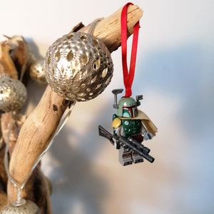 May include: A green and brown Lego Star Wars Boba Fett minifigure ornament with a red ribbon hanging from a branch. The minifigure is holding a black blaster rifle.