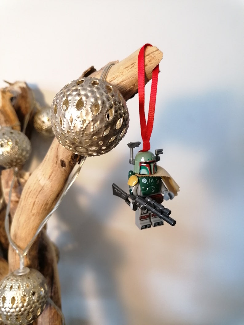 May include: A green and brown Lego Star Wars Boba Fett minifigure ornament with a red ribbon hanging from a branch. The minifigure is holding a black blaster rifle.