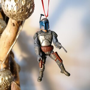 May include: A gray and blue Star Wars Boba Fett action figure ornament hanging from a red ribbon. The figure is wearing a helmet and holding a blaster.