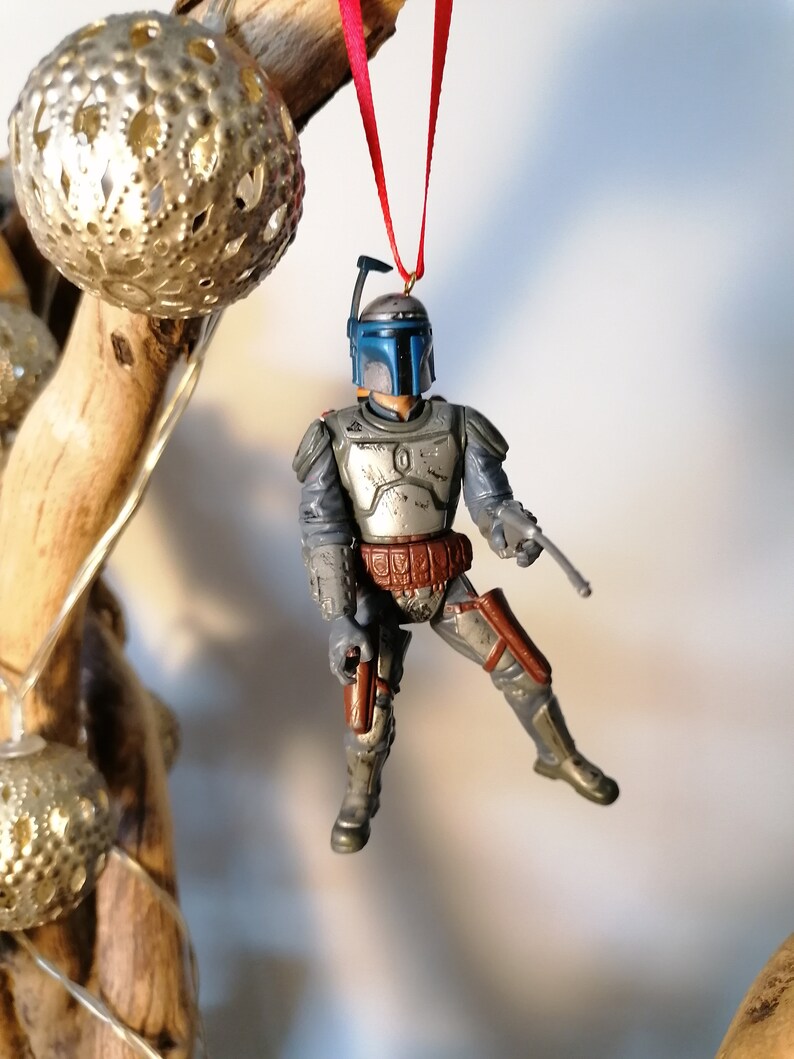 May include: A gray and blue Star Wars Boba Fett action figure ornament hanging from a red ribbon. The figure is wearing a helmet and holding a blaster.