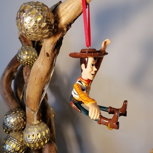 May include: A red string hangs from a branch with a plastic figurine of Woody from Toy Story. Woody is wearing his signature cowboy hat and vest. He is sitting with his legs outstretched and his arms raised.