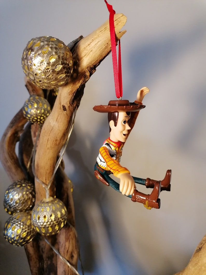May include: A red string hangs from a branch with a plastic figurine of Woody from Toy Story. Woody is wearing his signature cowboy hat and vest. He is sitting with his legs outstretched and his arms raised.