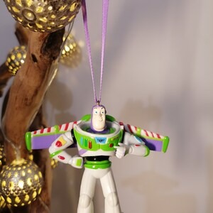 Peut inclure: Ornement de No&euml;l Buzz l'&Eacute;clair, le personnage de Toy Story. La figurine est principalement blanche, verte et violette, avec des d&eacute;tails rouges. Suspendu par un ruban violet. En arri&egrave;re-plan, des lumi&egrave;res d&eacute;coratives et une branche en bois.