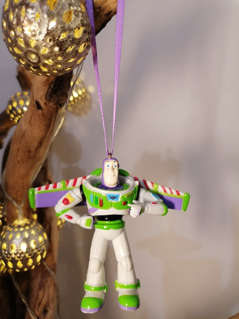 Peut inclure: Ornement de No&euml;l Buzz l'&Eacute;clair, le personnage de Toy Story. La figurine est principalement blanche, verte et violette, avec des d&eacute;tails rouges. Suspendu par un ruban violet. En arri&egrave;re-plan, des lumi&egrave;res d&eacute;coratives et une branche en bois.