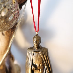 May include: A gold-colored ornament of a man wearing a cape and holding a sword. The ornament is hanging from a red ribbon.
