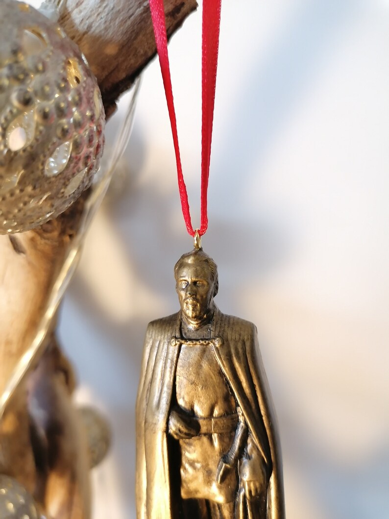 May include: A gold-colored ornament of a man wearing a cape and holding a sword. The ornament is hanging from a red ribbon.