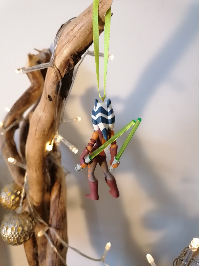 May include: A green and blue Star Wars character ornament with two green lightsabers. The ornament is hanging from a green ribbon.
