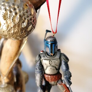 May include: A blue and gray Star Wars Boba Fett action figure ornament hanging from a red ribbon. The ornament is made of plastic and has a detailed design.