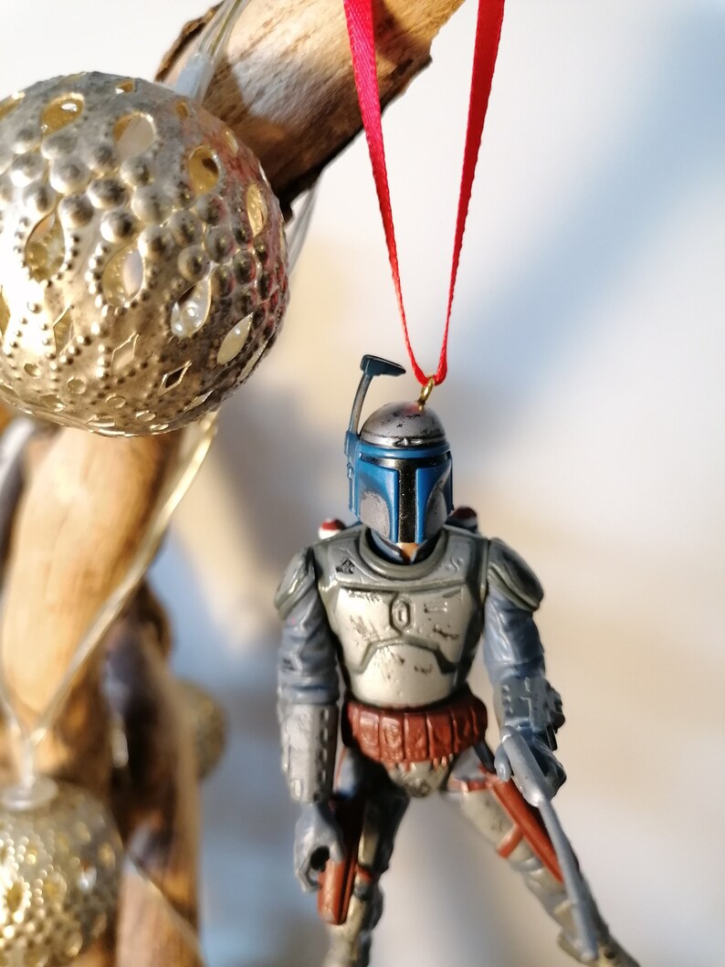 May include: A blue and gray Star Wars Boba Fett action figure ornament hanging from a red ribbon. The ornament is made of plastic and has a detailed design.
