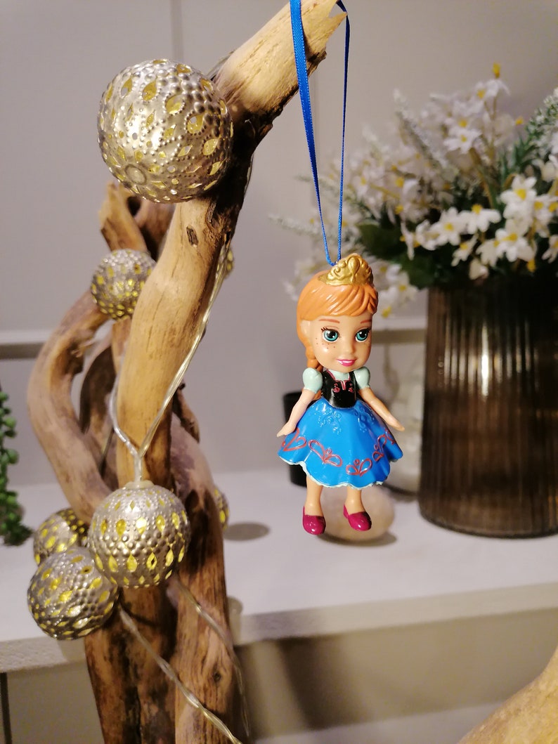 K&ouml;nnte beinhalten: Eine blaue und wei&szlig;e Puppe mit einer Krone, die an einem blauen Band h&auml;ngt. Die Puppe tr&auml;gt ein blaues Kleid mit roten Akzenten und hat braunes Haar und blaue Augen. Die Puppe h&auml;ngt an einem Ast mit goldfarbenen Ornamenten.