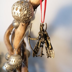 May include: A gold-coloured ornament of a winged creature with a bow and arrow. The ornament is hanging from a red ribbon.