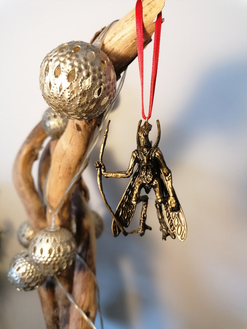 May include: A gold-coloured ornament of a winged creature with a bow and arrow. The ornament is hanging from a red ribbon.