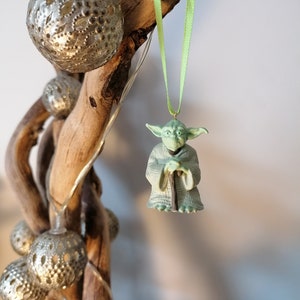 May include: A green Yoda Christmas ornament hanging from a green ribbon. The ornament is made of plastic and features Yoda in his signature pose, holding his staff.