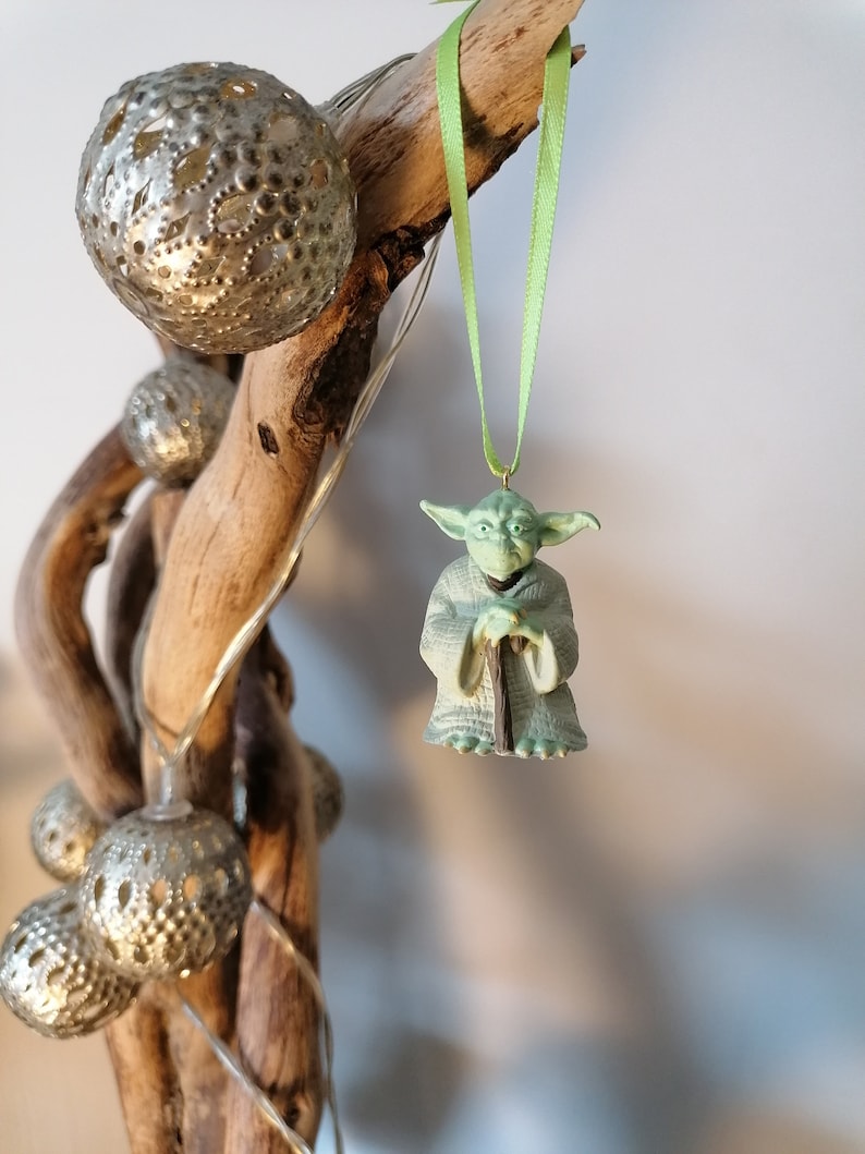 May include: A green Yoda Christmas ornament hanging from a green ribbon. The ornament is made of plastic and features Yoda in his signature pose, holding his staff.