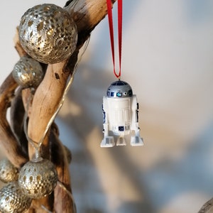 K&ouml;nnte beinhalten: Ein wei&szlig;er und blauer R2-D2 Star Wars Charakter-Anh&auml;nger, der an einem roten Band an einem Treibholzast mit silbernen Ornamenten h&auml;ngt.