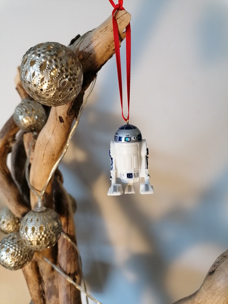 K&ouml;nnte beinhalten: Ein wei&szlig;er und blauer R2-D2 Star Wars Charakter-Anh&auml;nger, der an einem roten Band an einem Treibholzast mit silbernen Ornamenten h&auml;ngt.