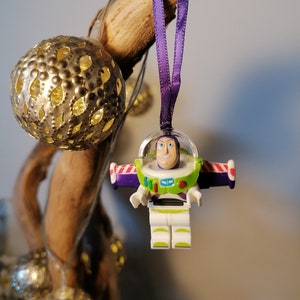 May include: A purple and white Christmas bauble featuring a plastic figurine of Buzz Lightyear from the Toy Story franchise. The bauble is hanging from a purple ribbon.